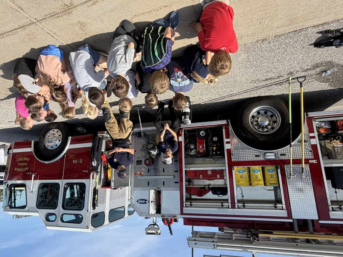 Students listen to firefighters near firetruck.